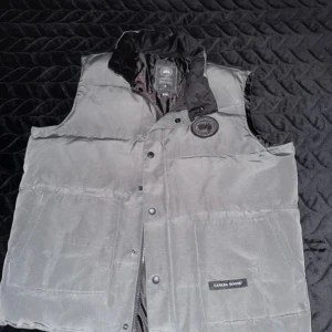 Grå dunväst från Canada Goose XL - Snygg grå dunväst från Canada Goose i storlek XL. Västen har hög krage, två stora fickor framtill och klassisk logotyp på bröstet. Stängs med både dragkedja och tryckknappar. Perfekt för kalla dagar och har en stilren, quiltad design.