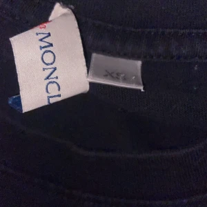 Svart Moncler t-shirt XS - Svart t-shirt från Moncler i storlek XS med klassisk rund hals och korta ärmar. På bröstet sitter Monclers ikoniska logotyp i vitt, blått och rött. T-shirten har en clean och stilren look, perfekt för dig som gillar exklusiva märken.