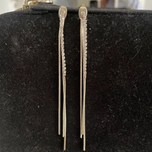 Långa silverörhängen med strass - Säljer ett par eleganta långa örhängen i silverfärg med smala kedjor och glittrande strass längs ena sidan. Perfekta för dig som gillar stilrena och trendiga smycken med lite extra bling.
