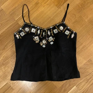 Ibiza topp - Svart linne med tunna axelband och snygga detaljer framtill i form av guldiga paljetter och pärlor i olika former. Toppen har en lätt glansig yta och justerbara axelband. Perfekt för dig som vill sticka ut med en unik och glittrig look.