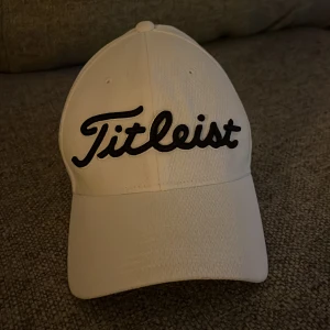 Vit Titleist keps Pro V1 - Snygg vit keps från Titleist med broderad svart logga framtill och texten Pro V1 på sidan. Kepsen har även FJ-logga och en patch bak. Tillverkad i ventilerande polyester, perfekt för golfbanan eller en sportig look.