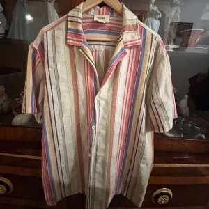 Randig skjorta från Pull & bear i linne - Säljer en färgglad randig skjorta från Pull & bear i linne. Skjortan har korta ärmar, knappar framtill och en avslappnad passform. Färgerna är blandade med inslag av blått, rött, grönt, beige och gult. Perfekt för varma dagar och chill vibes. 