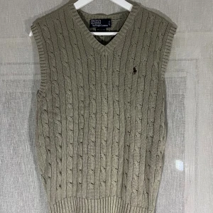 Beige stickad väst från Polo Ralph Lauren - Kabelstickad beige väst från Polo Ralph Lauren med v-ringning och klassisk broderad logga på bröstet. Västen har ribbade kanter och är tillverkad i mjuk bomull, perfekt för lager-på-lager-stil.