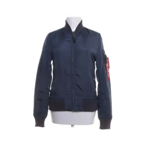 Mörkblå bomberjacka från Alpha Industries - Säljer en mörkblå bomberjacka från Alpha Industries med klassisk passform. Jackan har ribbade muddar, dragkedja framtill och en ikonisk röd tag på ärmen. Tillverkad i glansig polyester och har långärmad design. Perfekt för dig som vill ha en stilren och tidlös look.