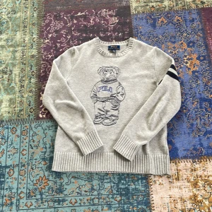 Grå stickad tröja Polo Ralph Lauren - Grå stickad tröja från Polo Ralph Lauren med ikonisk björn på bröstet och två mörkblå ränder på ena ärmen. Tröjan har rund hals, ribbade muddar och är långärmad. Perfekt för dig som gillar klassisk stil.