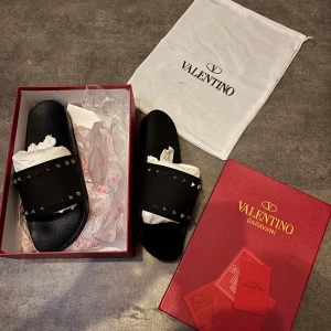 Valentino Garavani svarta sandaler - Svarta sandaler från Valentino Garavani med bred rem dekorerad med ikoniska nitar. Snygg och stilren design med platt sula och öppen tå. Tillverkade i Italien med exklusiv känsla och levereras med originalkartong och dustbag.