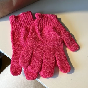 Rosa skrubbhandskar i syntetmaterial - Ett par knallrosa skrubbhandskar med grov yta, perfekta för exfoliering i duschen. De är tillverkade i syntetmaterial och har ribbade muddar för bra passform. Handskarna är flexibla och täcker hela handen med separata fingrar. Helt oanvända