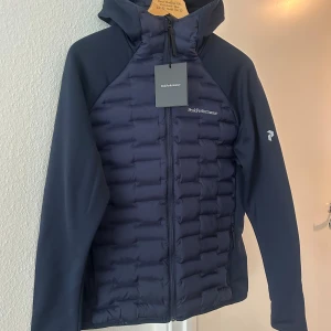 Marinblå Peak Performance jacka  - Mörkblå pufferjacka från Peak Performance med huva och hel dragkedja. Jackan har quiltad framsida och rygg, släta ärmar och diskret logga på bröstet och ärmen. Perfekt för kyliga dagar och har en sportig, stilren look. Helt ny med prislapp i storlek L