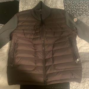 Grå Moncler pufferjacka med dragkedja - Snygg pufferjacka från Moncler i mörkgrått med quiltad front och släta ärmar. Jackan har hög krage, två dragkedjefickor och Moncler-logga på ärmen. Perfekt för kyliga dagar och ger en sportig vibe.