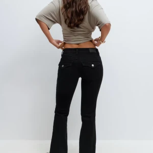 Svarta bootcut jeans från Gina - Svarta jeans med bootcut-snitt och snygga fickor med lock och silverknappar bak. Låg midja och klassisk femficksdesign. Jeansen har en stilren look och är tillverkade i stretchig denim för en bekväm passform.