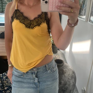 Gul linnetopp med svart spets Amisu - Säljer en gul linnetopp från Amisu med tunna axelband och snygg svart spetsdetalj längs v-ringningen. Toppen har en slät, mjuk yta och är perfekt för dig som gillar en trendig och feminin look. Passar till både jeans och kjol för en cool stil. Passar också storlek S.