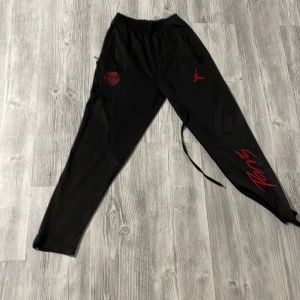 Svarta PSG x Jordan mjukisbyxor - Svarta mjukisbyxor från Jordan och PSG med röda detaljer, logga på vänster lår och Jumpman på höger. Tight passform med snörning i midjan och coolt mönstrade paneler längs benen. Perfekta för en sportig och street-inspirerad look.