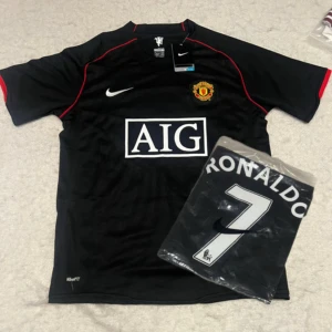 Manchester United Ronaldo 7 svart tröja - Säljer en svart Manchester United fotbollströja från Nike med AIG som sponsor på bröstet och klubbmärke på vänster sida. Tröjan har röda detaljer, korta ärmar och Ronaldo 7 tryckt på ryggen. Tillverkad i lätt och ventilerande material, perfekt för fotboll eller supporterliv.