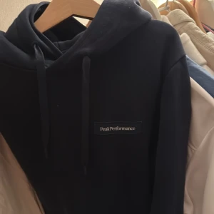 Svart hoodie Peak Performance S - Svart hoodie från Peak Performance i storlek S. Klassisk modell med huva och snörning, samt diskret logga på bröstet. Perfekt för chill dagar eller när du vill ha en enkel och snygg look.