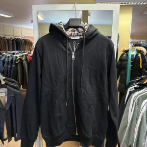 Svart hoodie från Burberry med dragkedja - Svart hoodie från Burberry med klassiskt rutigt foder i huvan och broderad logga på bröstet. Tröjan har hel dragkedja, snörning i huvan och två stora fickor framtill. Tillverkad i 100% bomull för en skön och mjuk känsla.