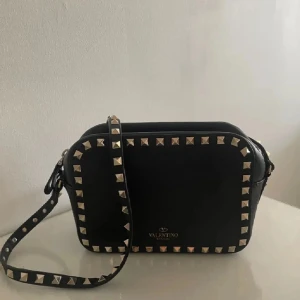 Svart valentino väska  - Snygg svart axelväska från Valentino med guldfärgade nitar runt kanterna och på axelremmen. Väskan är i fint skick men skadades lite på skinnet på sidan av bandet där bandet sitter fast i väskan, har lagat det hos en skräddare så nu är den som ny. Hör av dig vid frågor, pris kan diskuteras 