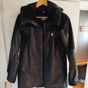 Svart och grå jacka från 8848 Altitude - Snygg svart och mörkgrå jacka från 8848 Altitude med huva och dragkedja framtill. Jackan har två sidofickor med dragkedja och en sportig look, perfekt för kyliga dagar. Märkeslogga på bröstet och praktiska detaljer för extra funktion.