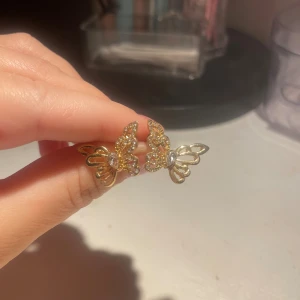Guldiga fjärilsörhängen med stenar - Säljer ett par snygga örhängen i form av fjärilar i guldton med glittrande stenar. De har en öppen och detaljrik design som verkligen fångar ögat. Perfekta för dig som vill ha något unikt och glammigt i smyckeslådan.