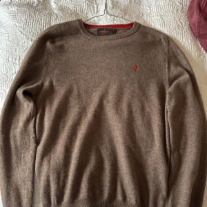 Brun merinoullströja från Morris - Säljer en brun tröja från Morris i 100% merinoull. Tröjan har rund halsringning, ribbade muddar och en broderad orange logga på bröstet. Perfekt för kyliga dagar och riktigt skön mot huden. Och sjukt snygga höst färger!