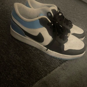 Nike Air Jordan 1 Low blå/vit/svart - Säljer ett par Nike Air Jordan 1 Low sneakers i blått, vitt och svart. De är i storlek 39, har tecken på använding, men annars bra skick. De är inte äkta.