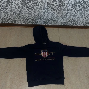 Marinblå hoodie från GANT - 	•	Märke: Gant 	•	Färg: Mörkblå (navy) 	•	Design: Huva med dragsnören, känguruficka framtill 	•	Detaljer: Broderad Gant-logga med sköld (röd-vit-randig) och texten “1949 GANT AMERICAN SPORTSWEAR” i guld 	•	Storlek: XS 	•	Stil: Klassisk och stilren hoodie i college-stil, perfekt för en preppy look eller avslappnad vardag