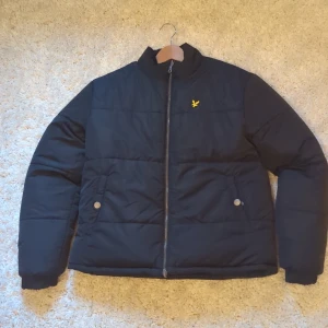 Vändbar Jacka Puffer Lyle & Scott - Säljer en svart pufferjacka från Lyle & Scott med gul broderad logga på bröstet. Jackan är vändbar med ena sidan helsvart och andra sidan mönstrad i blått och vitt. Dragkedja framtill och fickor med både knapp och dragkedja. Perfekt för kalla dagar!