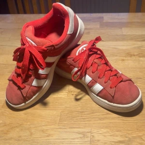 Adidas Campus röda sneakers mocka - Fräscha och trendiga sneakers i rött med de klassiska vita ränderna på sidorna. Skorna är tillverkade i ett slitstarkt material som ger både bra komfort och hållbarhet.  Skorna är i gott skick men har lite smuts som satt sig i tyget – detta märks mest på nära håll och inte på avstånd. Invändigt finns två små hål vid hälarna, men de påverkar inte användningen eller komforten.  Perfekta vardagsskor för dig som vill ha en snygg, sportig och tidlös look! 
