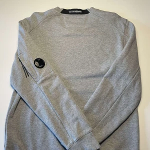 Grå sweatshirt från C.P. Company - En klassisk grå sweatshirt från CP company. En otroligt skön och snygg tröja med inga skavanker och är i bra skick. Det är inte alls mycket använd. Storlek L men sitter lite mindre. För mer info är det bara att skriva