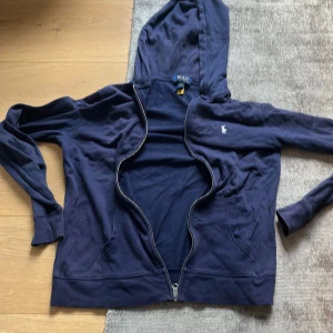 Marinblå hoodie från Polo Ralph Lauren - Snygg marinblå hoodie från Polo Ralph Lauren med dragkedja och klassisk broderad logga på bröstet. Tröjan har huva, långa ärmar och är tillverkad i mjuk bomull. Perfekt för en chill och stilren look. Pris kan diskuteras. 18-20 år är som storlek S