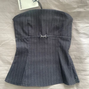 tubetop med bälte från Bershka - Aldrig använd och prislappen är kvar! En snygg mörkgrå tubetop från Bershka. Toppen är axelbandslös och har ett liten metallbälte framtill som detalj. Figurnära passform och lätt utsvängd nertill, perfekt festtopp som går att klä upp eller ner!