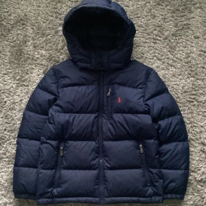 Mörkblå pufferjacka Polo Ralph Lauren - Snygg mörkblå pufferjacka från Polo Ralph Lauren med huva, tre dragkedjefickor och ikonisk röd logga på bröstet. Jackan har en quiltad design och stängs med dragkedja framtill. Perfekt för kalla dagar och riktigt stilren look.