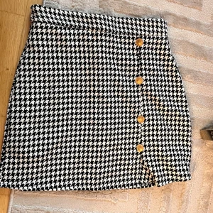 Rutig svartvit kortkjol med knappar - Snygg svartvit kortkjol med klassiskt rutigt hundtandsmönster och dekorativa guldknappar framtill. Kjolen har en rak passform och är perfekt för dig som gillar en stilren och trendig look.