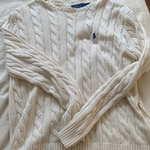 Vit kabelstickad tröja Polo Ralph Lauren - Säljer en klassisk vit kabelstickad tröja från Polo Ralph Lauren i storlek M. Tröjan har rund hals, långärmade ribbade muddar och den ikoniska broderade loggan på bröstet. Perfekt för dig som gillar stilrena och tidlösa plagg.