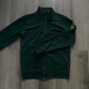 Mörkgrön ziptröja från Stone Island - Snygg mörkgrön ziptröja från Stone Island med klassisk patch på ärmen. Tröjan har ståkrage, hel dragkedja och två fickor fram. Tillverkad i mjuk bomull som är skön att ha på sig. Perfekt för dig som gillar streetwear och vill ha något stilrent.