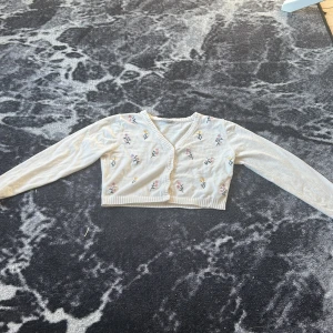 Vit kofta med broderade blommor - Superfin vit kofta med broderade blommor i rosa, lila och gula. Croppad modell med v-ringning och knappar framtill. Långärmad och ribbade muddar. Perfekt för vår och sommar när du vill ha något lätt och gulligt.