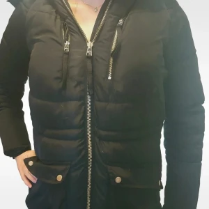 Svart Jacka med fuskpäls från Saint Tropez - Snygg svart vinterjacka från Saint Tropez med dragkedja framtill, två fickor med knappar och huva med fluffig fuskpäls i beige. Perfekt för kalla dagar och ger en cool, femenin look med sina detaljer och quiltade partier.