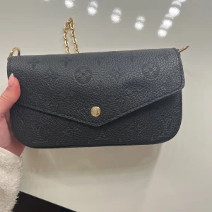 Svart märkes väska  - Snygg svart axelväska i präglat skinn med klassiskt monogrammönster. Väskan har guldfärgad kedja och tryckknapp framtill. Perfekt storlek för det viktigaste och ger en lyxig känsla till din outfit.