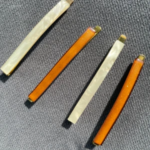 Set med fyra raka hårspännen - Fyra stilrena hårspännen i rektangulär form, två i skimrande pärlemovit och två i glittrande bärnstensbrun färg. Baksidan är i guldtonad metall. Perfekta för att ge en snygg touch till din frisyr.