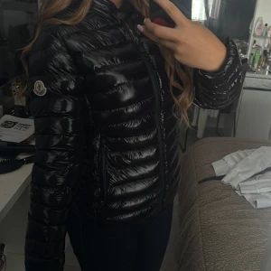 Svart dunjacka från Moncler - Säljer en svart, glansig dunjacka från Moncler med klassisk logga på ärmen. Jackan har quiltad design, dragkedja framtill och hög krage. Perfekt för kalla dagar och ger en clean, stilren look. Passformen är normal och jackan är långärmad.