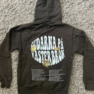 Hov1 merch hoodie - Hov1 merch hoodie med  'Gudarna på Västerbron' och turnélista på ryggen. Snygga små tryck med blommor, bil och krona. Liten broderad krona på framsidan. Klassisk huva och muddar. Perfekt för dig som gillar bandmerch och streetstyle.