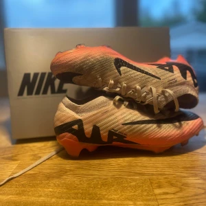 Nike Air Zoom fotbollsskor beige/rosa - Säljer ett par Nike Air Zoom fotbollsskor i beige och rosa med svarta detaljer och stora Nike-loggor på sidorna. Skorna har snörning och en lätt, stickad ovandel i syntetmaterial samt rosa dobbar för bra grepp på planen. Perfekta för dig som vill sticka ut på matchen.