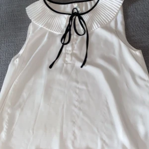 Vit gullig blus  Zara - Supersöt vit  blus från Zara med plisserad krage och svart knytband vid halsen. Blusen har knappar framtill och är gjord i ett lätt, slätt material som känns lyxigt. Perfekt för dig som gillar en chic och stilren look.            Den har några småfläckar framtill men som knappt syns  (titta på bild två) , annars superbra skick! 