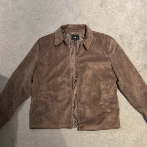 Brun mockajacka från H&M - Snygg brun mockajacka från H&M med klassisk krage och dragkedja framtill. Jackan har två diskreta sidfickor och är i ett mjukt material som ger en stilren och clean look. Perfekt för dig som gillar minimalistisk stil och vill ha något som sticker ut utan att vara för mycket.