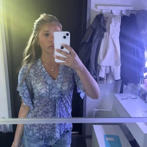 Blommig blå blus med volangärm - Säljer en söt blommig blus i ljusblått med v-ringning och volangärmar. Blusen har ett fint knyt framtill och är gjord i ett lätt, transparent material som känns luftigt. Perfekt för dig som gillar romantiska detaljer och en feminin stil.