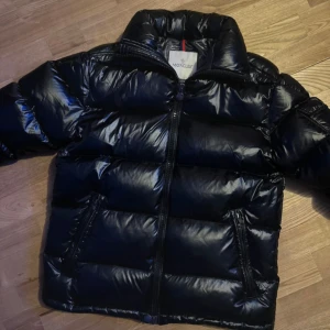 Svart Moncler pufferjacka - Snygg svart pufferjacka från Moncler med glansig finish och klassisk logga på ärmen. Jackan har hög krage, dragkedja framtill och flera praktiska fickor. Perfekt för kalla dagar när du vill hålla stilen och samtidigt vara varm.