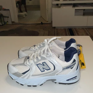New Balance 530 vita och blå sneakers - Snygga New Balance 530 sneakers i vitt och marinblått med silverdetaljer. Skorna har mesh och syntetmaterial, klassisk snörning och en chunky sula med Abzorb-dämpning. Perfekta för dig som gillar retrovibbar och sportig stil.