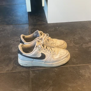 Nike Air Force 1 vita sneakers - Klassiska Nike Air Force 1 sneakers i vitt skinn med svart swoosh och svart detalj på hälen. Lågt skaft, perforerad tå och snörning framtill. Perfekta för dig som gillar streetstyle och tidlös design.