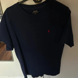 Marinblå t-shirt från Polo Ralph Lauren - Snygg marinblå t-shirt från Polo Ralph Lauren i storlek L. Klassisk design med korta ärmar och rund hals. T-shirten har det ikoniska röda Polo-logot på bröstet och är gjord i mjuk bomull för skön komfort.