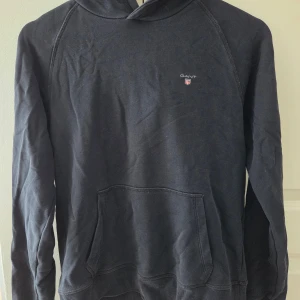 Svart hoodie från GANT med huva - Svart hoodie från GANT med klassisk huva och magficka. Diskret GANT-logga broderad på bröstet. Perfekt för chill dagar eller när du vill ha en enkel och stilren look. Tillverkad i mjuk bomull för skön känsla.