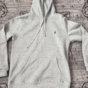 Grå hoodie från Polo Ralph Lauren - Snygg ljusgrå hoodie från Polo Ralph Lauren med klassisk broderad logga på bröstet. Tröjan har huva med snörning och en stor magficka. Tillverkad i mjuk bomullsmix, perfekt för chill dagar. Enkel och stilren design som funkar året runt.
